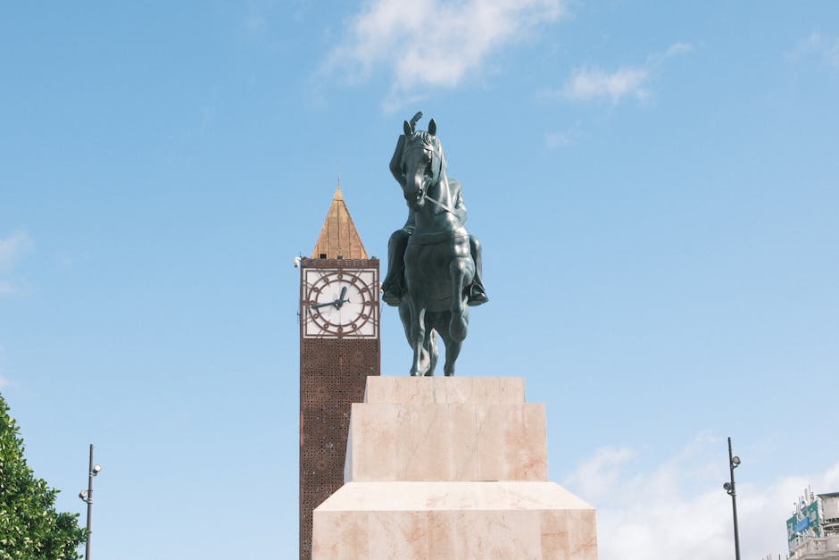 A bronze equestrian statue featuring a rider on horseback is positioned on a large stone pedestal in front of a historic clock tower with a pointed, golden-brown spire. The clock displays a time of ap