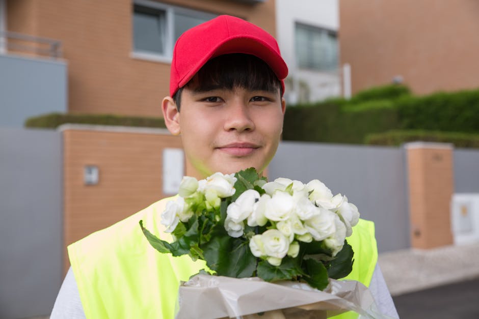 A young male flower delivery courier wearing a red cap and high-visibility yellow vest is outdoors, holding a bouquet of fresh white roses wrapped in brown paper and clear cellophane. The roses have f