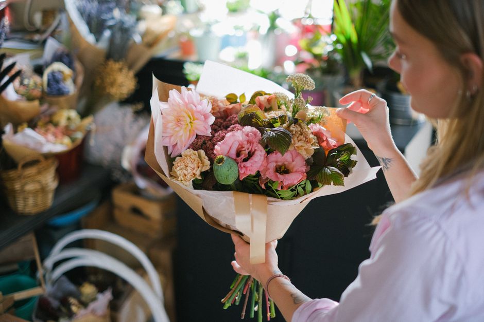 A woman with light brown hair is holding a bouquet of fresh flowers wrapped in white paper and tied with a beige ribbon, featuring a variety of pink flowers including a large dahlia, roses, and smalle