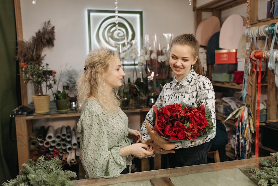 A young woman with light brown hair tied back in a ponytail, wearing a patterned blouse, is holding a vibrant bouquet of red roses, lilies, and other mixed flowers wrapped in green foliage and brown p